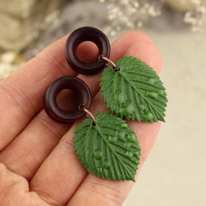 May include: A pair of dark brown wooden earrings with a circular design, each connected to a green leaf-shaped charm with water droplet details. The earrings are held in a hand.