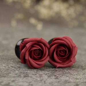 May include: Two dark red rose-shaped plugs with a black base, set against a grey background. The rose petals are detailed and textured, creating a realistic appearance. These plugs are likely for ear piercings, and are a decorative accessory.