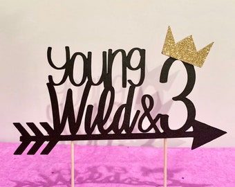 以下が含まれることがあります： 「young wild & 3」という文字と右を向いた矢印が書かれた黒いケーキトッパー。3の上に金色のグリッターの王冠が置かれている。