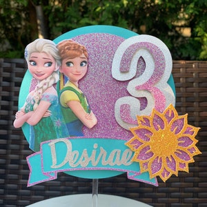 Könnte beinhalten: Ein türkisfarbener und rosafarbener Tortenaufsatz mit einer weißen Zahl 3, einer Sonnenblume und zwei Zeichentrickfiguren aus dem Film Frozen. Der Aufsatz enthält auch den Namen "Desiree" in silbernen Glitzer.
