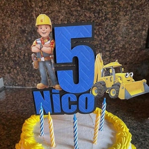 May include: A white cake with yellow frosting and blue and yellow candles. The cake is decorated with a blue number 5, the name "Nico", and a cartoon character from the show "Bob the Builder".