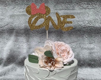 May include: A white cake with a gold glitter "one" and a pink bow on top. The cake is decorated with white and pink flowers.