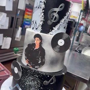 May include: A multi-tiered cake with a music theme. The cake features black, white, and silver accents, including musical notes, treble clefs, and vinyl record decorations. A picture of Michael Jackson is on the cake. The top of the cake has the name "Javin" on it.