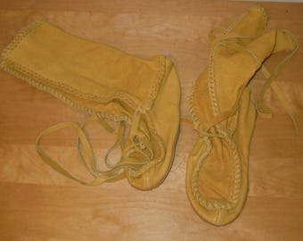 May include: A pair of mustard yellow soft leather moccasin boots featuring a high-top design with visible whipstitch detailing along the seams and long leather laces for tying around the ankle or calf for a secure fit.