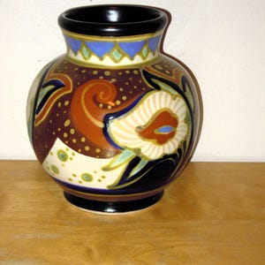 May include: A decorative ceramic vase with a black rim and base. The vase features a floral design with brown, orange, blue, and white accents. The vase is approximately 15 cm tall.