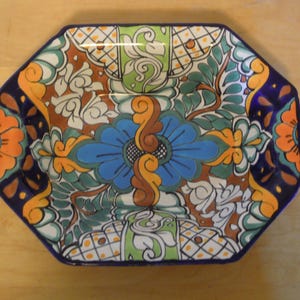 May include: An octagonal ceramic plate with a vibrant floral design. The plate features a blue border and is adorned with a central blue flower, orange accents, and green foliage. The intricate pattern is set against a white background.