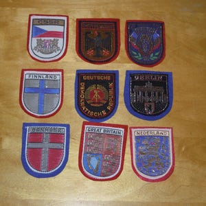 Peut inclure: Collection de neuf écussons brodés en forme de bouclier, chacun avec un design différent. Les écussons présentent des drapeaux, des armoiries et du texte, dont "CSSR", "Deutschland", "Finnland", "Berlin", "Danmark", "Great Britain" et "Nederland".