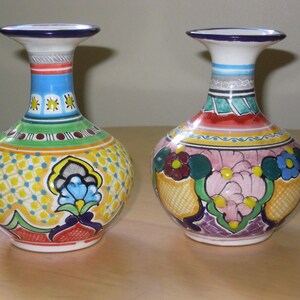 May include: Two hand-painted ceramic vases with a bulbous shape and flared necks. The vases feature vibrant colours, including blue, yellow, green, and red, with floral and geometric patterns. Each vase is approximately 15 cm tall.