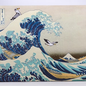 Peut inclure: Impression sur toile de "La Grande Vague de Kanagawa" de Hokusai, avec des mouettes de dessin animé. L'œuvre représente une grande vague océanique avec des bateaux et le Mont Fuji en arrière-plan. La palette de couleurs comprend des nuances de bleu, de blanc et de beige.