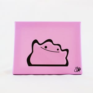 May include: A pink square canvas featuring a black outline of a Ditto character with a smiling face. The canvas has a solid pink background and a black border around the character. A small signature is in the bottom right corner.