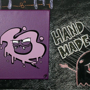 May include: Purple canvas painting with a cartoon character in a pink blob shape with a toothy grin. The background is purple and the text "HAND MADE" is written in white on a black background.