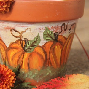 May include: A terracotta-colored flower pot with a hand-painted pumpkin design. The pumpkins are orange with green leaves and sit on a bed of green grass. The pot is decorated with orange flowers.