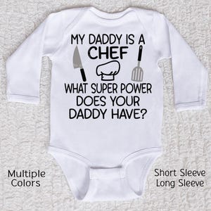 May include: White baby bodysuit with long sleeves. The text "MY DADDY IS A CHEF" is printed in black, along with a chef's hat, knife, and spatula graphic. The text "WHAT SUPER POWER DOES YOUR DADDY HAVE?" is also printed on the bodysuit.