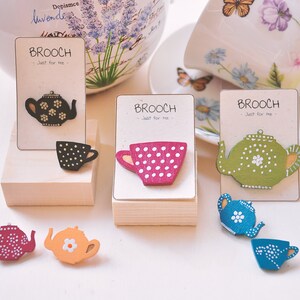 May include: A set of six wooden brooches in the shape of teapots and teacups. The brooches are painted in various colors, including black, white, pink, green, and blue. The brooches are displayed on small wooden stands with the text "Brooch - Just for me" printed on cards.