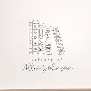 May include: An open book with a black ink stamp on the cream-colored page. The stamp features a line drawing of four books standing upright with botanical details on their spines, and the text "library of Allie Johnson" printed below.
