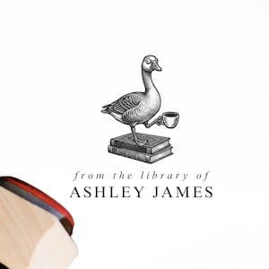 May include: A wooden rubber stamp with a black handle and a light wood base, positioned next to a book page featuring a black ink illustration of a goose standing on a stack of books holding a cup, with the text from the library of ASHLEY JAMES.