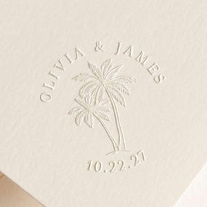 May include: An off-white card with a blind embossed design featuring two palm trees in the center. The text OLIVIA & JAMES is arched above the trees, and the date 10.22.27 is printed in a curved line below the palm trees on the textured paper.