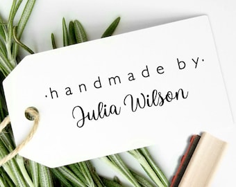 May include: A white rectangular paper tag with a hole and a jute string, featuring the black printed text "handmade by." and "Julia Wilson" in a script font. The tag is positioned over green rosemary sprigs next to a wooden rubber stamp.