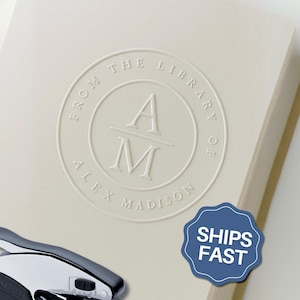 May include: A chrome and black metal embosser tool positioned next to a cream-colored paper sheet featuring a circular embossed design. The design includes the text FROM THE LIBRARY OF ALEX MADISON surrounding the centered initials A and M.