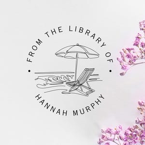 May include: A circular black ink stamp design on a white surface featuring a line art illustration of a beach umbrella and a lounge chair by the ocean. The text reads FROM THE LIBRARY OF and HANNAH MURPHY in a clean, sans-serif typeface.
