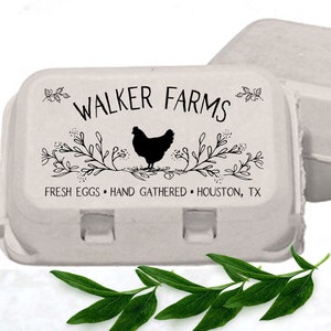 May include: Close-up view of two light gray cardboard egg cartons.  The carton in the foreground features a black and white design with the text 'WALKER FARMS', 'FRESH EGGS', 'HAND GATHERED', and 'HOUSTON, TX'. A black silhouette of a chicken is centered within a decorative floral branch design. A sprig of green leaves rests in front of the cartons.