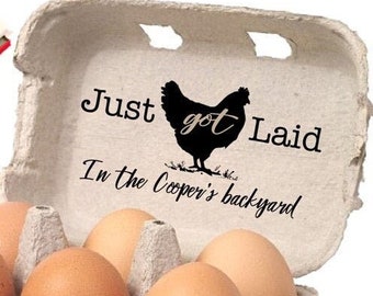 May include: A light brown molded pulp egg carton containing six brown eggs. The inside lid features a black silhouette of a chicken and the text "Just got Laid In the Cooper's backyard". A wooden rubber stamp is visible in the upper left corner.