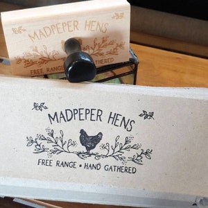 May include: A rectangular wooden rubber stamp with a black handle, featuring the text MADPEPER HENS, a hen illustration, and the words FREE RANGE HAND GATHERED. Below it, a light gray egg carton displays the same black ink stamped design.