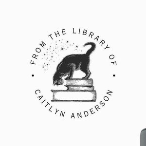May include: A black self-inking stamp with the text Trodat Printy 4924 and a red button. The stamp impression features a circular design with a cat standing on a stack of books, surrounded by stars and the text FROM THE LIBRARY OF CAITLYN ANDERSON.