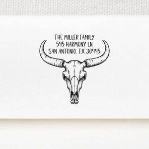 May include: A black self-inking rubber stamp positioned below a white envelope. The envelope features a black ink imprint of a longhorn steer skull graphic centered above the text THE MILLER FAMILY 545 HARMONY LN SAN ANTONIO TX 30445.