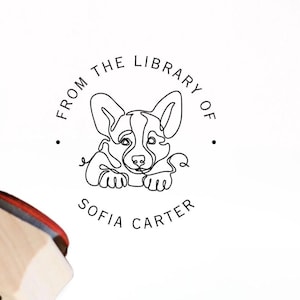 May include: A wooden stamp with a black handle and a red ink pad is next to an open book. The stamp has a circular design with a corgi illustration and the text "FROM THE LIBRARY OF SOFIA CARTER."