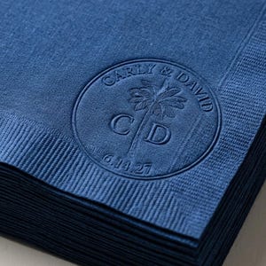 May include: A stack of deep blue paper napkins featuring a circular embossed design. The center displays a palm tree graphic with the letters C and D, surrounded by the text Carly & David and the date 6.14.27 in a circular border.