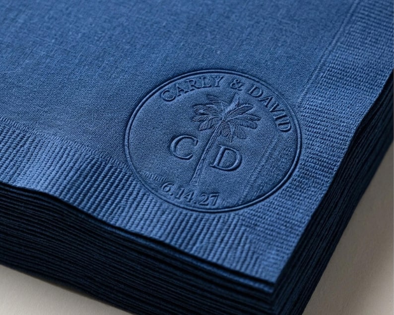 May include: A stack of deep blue paper napkins featuring a circular embossed design. The center displays a palm tree graphic with the letters C and D, surrounded by the text Carly & David and the date 6.14.27 in a circular border.