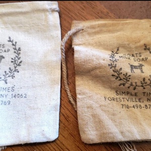 May include: Two rectangular drawstring pouches made of light-colored fabric. Each pouch features a printed design of a goat inside a wreath with the text GOATS MILK SOAP, SIMPLE TIMES, FORESTVILLE, NY 14062, and the phone number 716-498-8769.