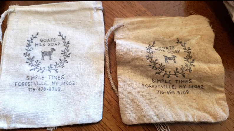 May include: Two rectangular drawstring pouches made of light-colored fabric. Each pouch features a printed design of a goat inside a wreath with the text GOATS MILK SOAP, SIMPLE TIMES, FORESTVILLE, NY 14062, and the phone number 716-498-8769.