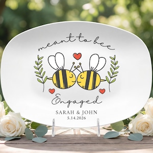 May include: An oval white ceramic platter featuring a graphic of two yellow and black bees with white wings, three red hearts, and green leafy sprigs. The text reads "meant to bee", "Engaged", "SARAH & JOHN", and the date "5.14.2026" in black.