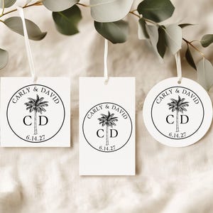 May include: Three white paper tags with white ribbon loops, featuring a black circular design with a palm tree illustration, the names Carly & David, the initials C D, and the date 6.14.27. The set includes one square, one rectangular, and one round tag.