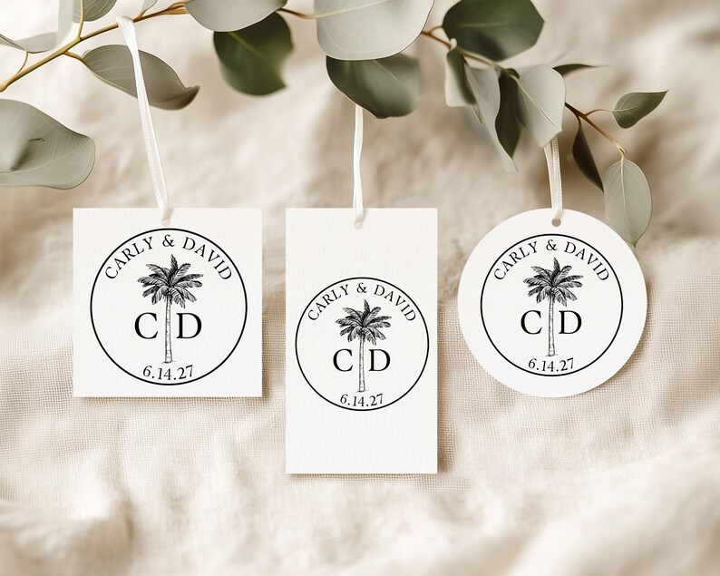 May include: Three white paper tags with white ribbon loops, featuring a black circular design with a palm tree illustration, the names Carly & David, the initials C D, and the date 6.14.27. The set includes one square, one rectangular, and one round tag.