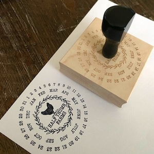 May include: A wooden stamp with a circular design featuring a rooster, a wreath of leaves, and the text "Half Thumb Farms". The stamp also includes a calendar with the months of the year and the numbers 1 through 31.