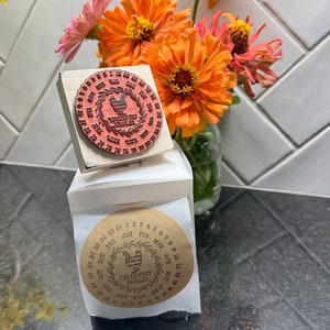 May include: A set of two round wooden stamps featuring a rooster design. The stamps are used to mark dates on parcels or other items. The larger stamp has the text "Rooster Creek Farms" and the smaller stamp has the text "Oh Glory Farms".