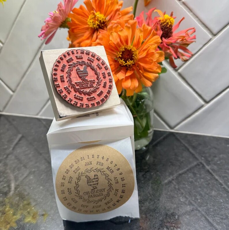 May include: A set of two round wooden stamps featuring a rooster design. The stamps are used to mark dates on parcels or other items. The larger stamp has the text "Rooster Creek Farms" and the smaller stamp has the text "Oh Glory Farms".