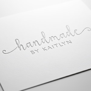 May include: A white card with the embossed text "handmade" in a cursive font, followed by "BY KAITLYN" in a smaller, sans-serif font. The card has a textured surface and is angled in the image.