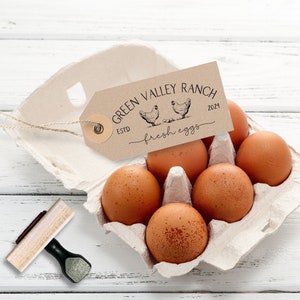 Peut inclure: Une bo&icirc;te &agrave; &oelig;ufs en carton contenant six &oelig;ufs bruns avec une &eacute;tiquette en papier beige attach&eacute;e par une ficelle. L'&eacute;tiquette porte le texte "GREEN VALLEY RANCH ESTD 2024 fresh eggs" et une illustration de deux poules. Un tampon en bois est pos&eacute; &agrave; c&ocirc;t&eacute;.
