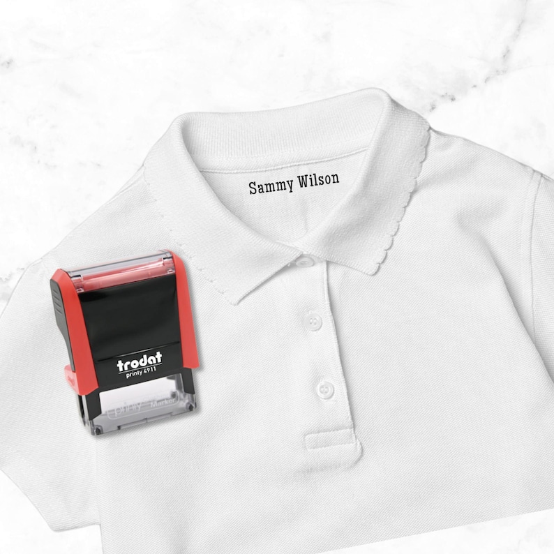 May include: A red and black Trodat stamp next to a white polo shirt. The shirt has the name "Sammy Wilson" printed on the collar. The stamp is a rectangular shape with the brand name "trodat printy 4911" printed on it.
