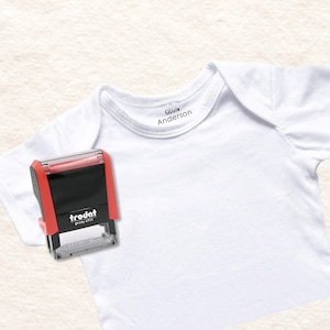 May include: A white baby bodysuit with the name "Olivia Anderson" printed on the collar. A red and black Trodat Printy 4911 stamp is positioned next to the bodysuit, for labelling clothing.