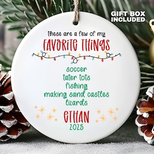 May include: A round white ceramic ornament featuring the text "these are a few of my FAVORITE THINGS" in black and red, followed by a list of green words: soccer, tater tots, fishing, making sand castles, lizards, the name ETHAN, and 2025.