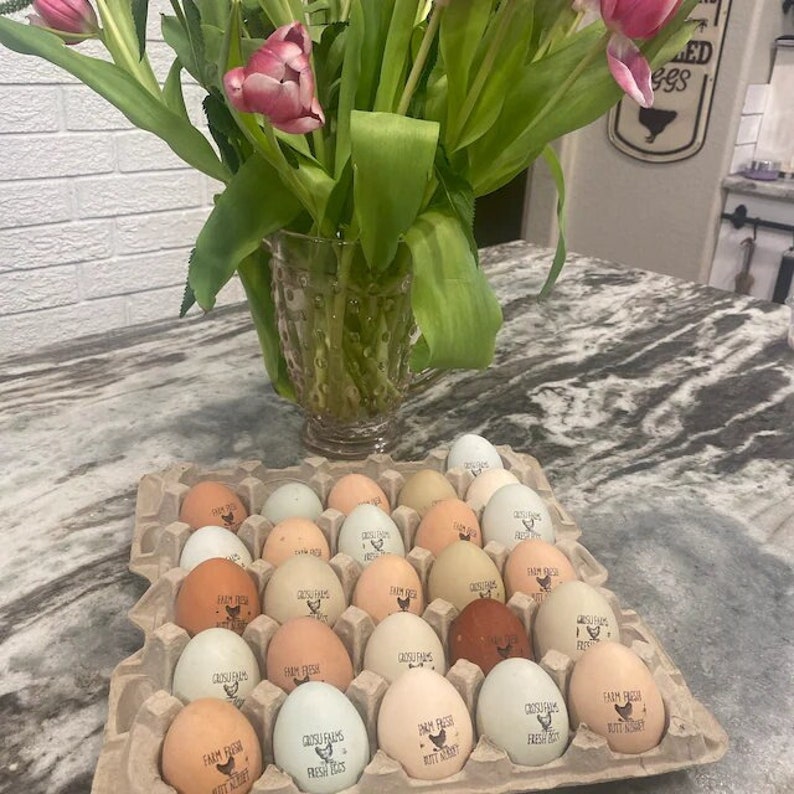 May include: A full carton of fresh eggs in various shades of brown, white, and blue-green, each stamped with "Farm Fresh" and a logo. A vase of pink tulips sits behind the carton, adding a touch of color to the scene.