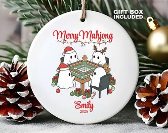 May include: A round white ceramic ornament featuring a graphic of four ghosts playing mahjong at a table. The text reads Merry Mahjong, Emily, and 2025. The ghosts wear holiday accessories like a Santa hat and reindeer antlers, surrounded by gifts.