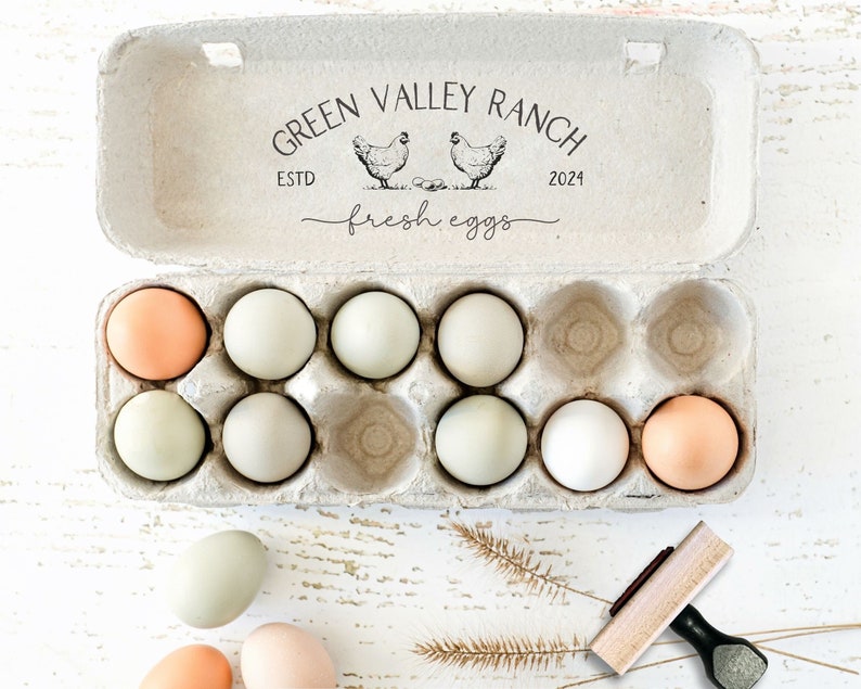 Peut inclure: Une bo&icirc;te &agrave; &oelig;ufs en p&acirc;te &agrave; papier grise contenant dix &oelig;ufs dans des tons vari&eacute;s de blanc, cr&egrave;me et brun clair. Le couvercle pr&eacute;sente un texte imprim&eacute; en noir indiquant GREEN VALLEY RANCH ESTD 2024 et fresh eggs avec deux illustrations de poules.
