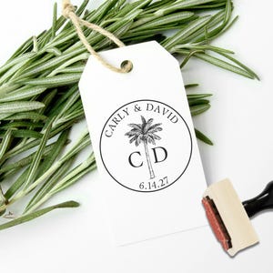 May include: A white rectangular paper gift tag with a jute string, featuring a circular black ink stamp design of a palm tree, the names Carly & David, the initials C D, and the date 6.14.27. A wooden rubber stamp with a black handle is nearby.