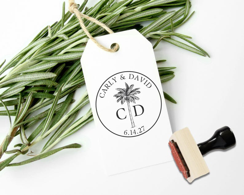 May include: A white rectangular paper gift tag with a jute string, featuring a circular black ink stamp design of a palm tree, the names Carly & David, the initials C D, and the date 6.14.27. A wooden rubber stamp with a black handle is nearby.